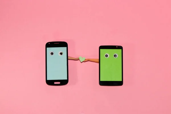 sending a love message mobile phones look at each other and one phone gives the heart to the other from hand to hand, both phones with copy space, pink background, creative modern design