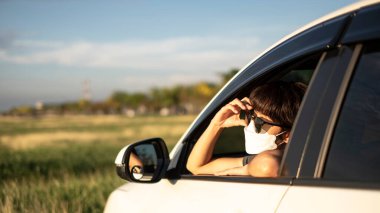 COVID-19 'un yayılmasını önlemek için maske takan kısa saçlı Asyalı turist kadın, New Normal turizm konseptinde doğaya göz kulak olmak için bir arabada oturuyor ve açık havaya çağrı yapıyor.