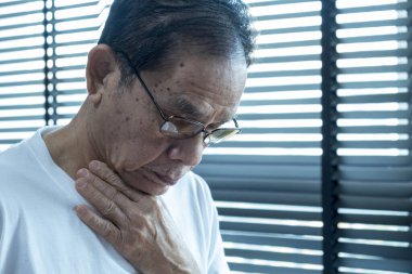 Asyalı kıdemli bir adam gözlük takarak öksürüyor, sabah uyandığında, yatak odasında, beyaz bir gömlek giyiyor, elleri boynundaydı.