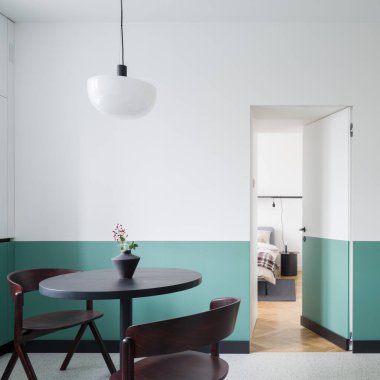 Modern tasarım yemek odası, koyu renk masa, iki sandalye şık lamba altında, yarı beyaz ve yeşil duvarlar ve yatak odasına açık kapılar.