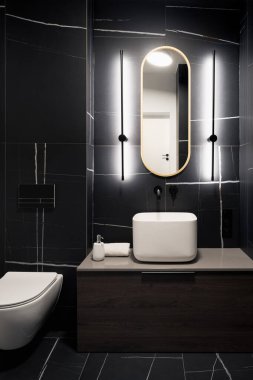 Siyah fayanslı koyu renkli banyo, modern kare lavabo ve kurşun ışıklı oval ayna.