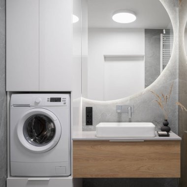 Modern ve işlevsel banyo, büyük yuvarlak ayna ve çamaşır makinesi.