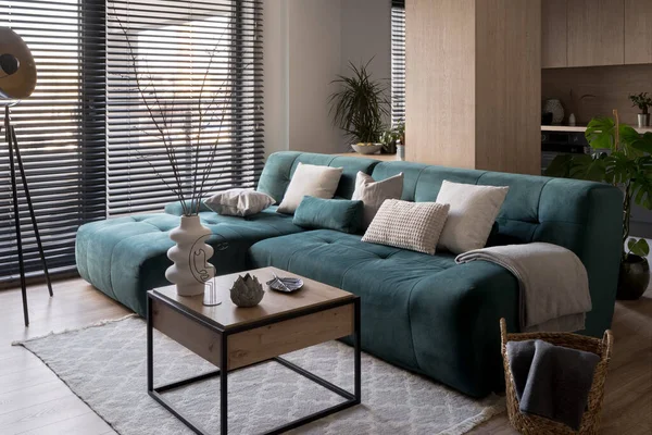 Cozy, blue corner sofa with square, wooden coffee table with decorations in modern living room with big windows with blinds