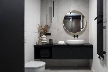 Modern tasarlanmış banyo dekoratif beton duvarlı, yuvarlak ayna siyah dolapta ve tuvalette lavabonun üzerinde.