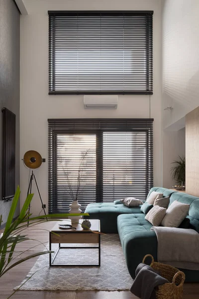 Big windows with blinds in lofty living room with cozy sofa, stylish decorations and simple coffee table