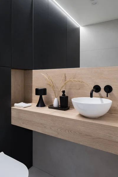 Simple and elegant bathroom with black furniture, wooden shelf, small white washbasin with black tap and mirror