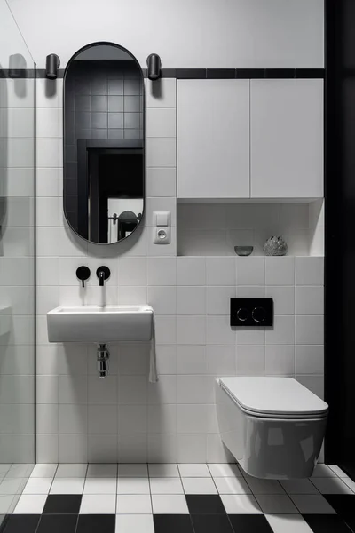 黒と白の床と壁のタイルで近代的なバスルーム 小さな洗面台と正方形のトイレの上に楕円形のミラーと — ストック写真