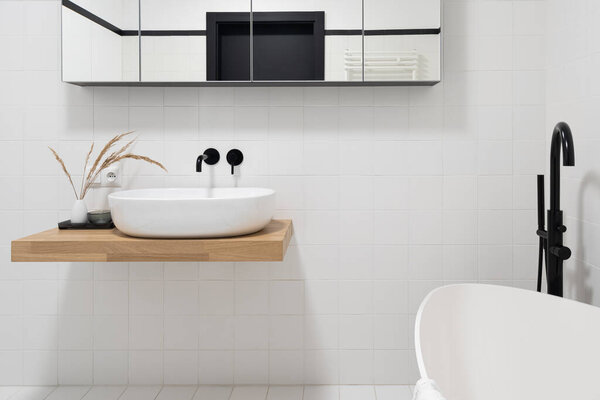 Small, white washbasin on wooden shelf in minimalist bathroom with white tiles and black taps