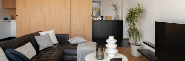 Panorama of stylish living room with leather, black corner sofa, simple coffee table, big tv and wooden, sliding doors
