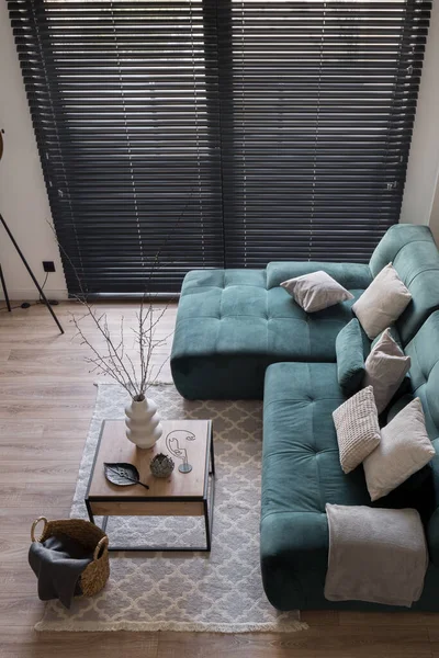 Top view of living room with big window with black blinds and big, cozy corner sofa with pillows