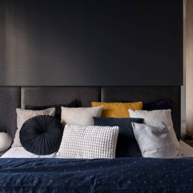 Modern karanlık yatak odasında yatak başlığı ve dekoratif yastıkları olan rahat bir yatağa yakın çekim.
