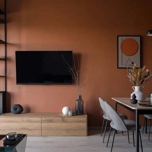 Tv screen on modern orange wall in stylish dining room with table, art and decorations