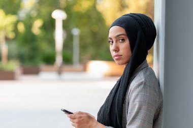 Front side view of a muslim adult woman leaning on a beam column looking at camera while using her phone in a park.