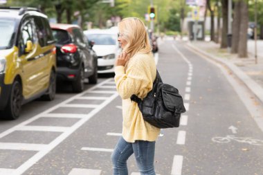 Blonde woman with a yellow sweater and a black bag crossing the street while touching her hair