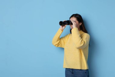 Dürbünle bakan Asyalı bir kadının mavi arka planlı stüdyo fotoğrafı.