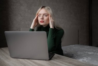 Yeşil golflü üzgün ve çok yorgun sarışın bir kadın evde dizüstü bilgisayarla çalışmaktan sıkıldı. Odada kötü bir ruh halinde masada oturan bir kız. Çevrimiçi öğrenme. Görünmez video konferansı.