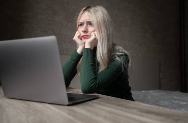 Yeşil golflü üzgün ve çok yorgun sarışın bir kadın evde dizüstü bilgisayarla çalışmaktan sıkıldı. Odada kötü bir ruh halinde masada oturan bir kız. Çevrimiçi öğrenme. Görünmez video konferansı.