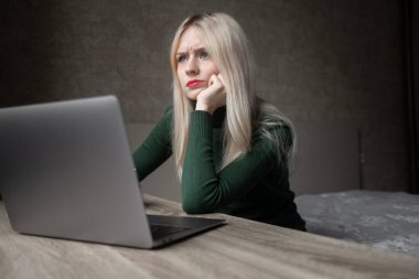 Yeşil golflü üzgün ve çok yorgun sarışın bir kadın evde dizüstü bilgisayarla çalışmaktan sıkıldı. Odada kötü bir ruh halinde masada oturan bir kız. Çevrimiçi öğrenme. Görünmez video konferansı.