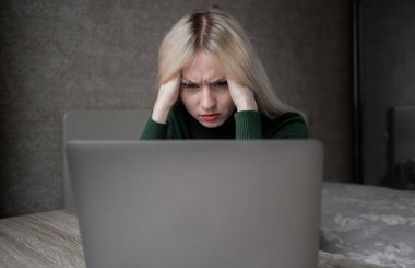 Yeşil golflü üzgün ve çok yorgun sarışın bir kadın evde dizüstü bilgisayarla çalışmaktan sıkıldı. Odada kötü bir ruh halinde masada oturan bir kız. Çevrimiçi öğrenme. Görünmez video konferansı.