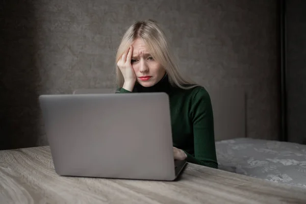 Uma mulher loira triste e muito cansada em um golfe verde está entediada trabalhando em casa com um laptop. Menina sentada à mesa de mau humor na sala. Aprendizagem online. Videoconferência feia. — Fotografia de Stock