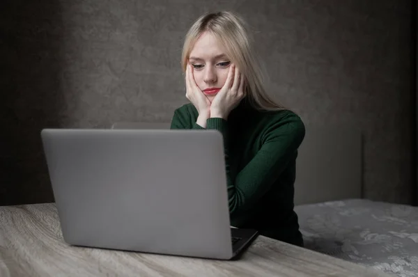 Een trieste en erg vermoeide blonde vrouw in een groene golf verveelt zich thuis te werken met een laptop. Zittend aan tafel in een slechte bui in de kamer. Online leren. Onooglijke videoconferentie. — Stockfoto