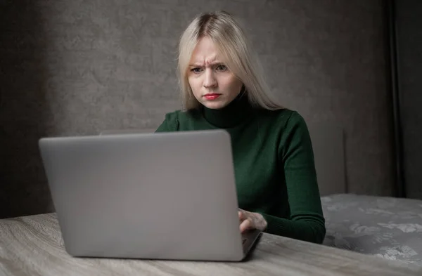 Een trieste en erg vermoeide blonde vrouw in een groene golf verveelt zich thuis te werken met een laptop. Meisje zit aan tafel in een slechte bui in de kamer. Online leren. Onooglijke videoconferentie. — Stockfoto