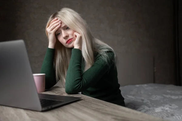 Een trieste en erg vermoeide blonde vrouw in een groene golf verveelt zich thuis te werken met een laptop. Meisje zit aan tafel in een slechte bui in de kamer. Online leren. Onooglijke videoconferentie. — Stockfoto