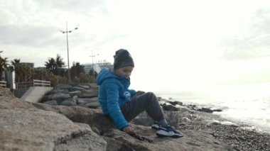 Okul çağındaki bir çocuk taşların üzerine oturur ve denize ya da okyanusa çakıl taşları atar. Sonbahar ya da ilkbahar. Sahil. Dinlenme ve faaliyet.