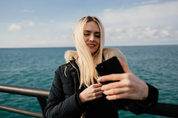 Güzel, gülümseyen sarışın bir kız telefonda denizin arka planında selfie çekiyor..