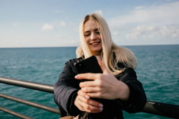 Güzel, gülümseyen sarışın bir kız telefonda denizin arka planında selfie çekiyor..