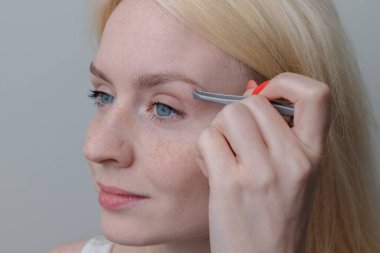 A beautiful young blonde girl with freckles plucks her eyebrows with tweezers. Natural beauty.