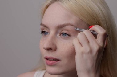 A beautiful young blonde girl with freckles plucks her eyebrows with tweezers. Natural beauty.