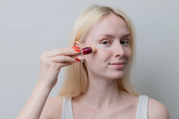  A young blonde woman with freckles applies concealer on her face under her eyes and masks circles under her eyes. Natural makeup. Text