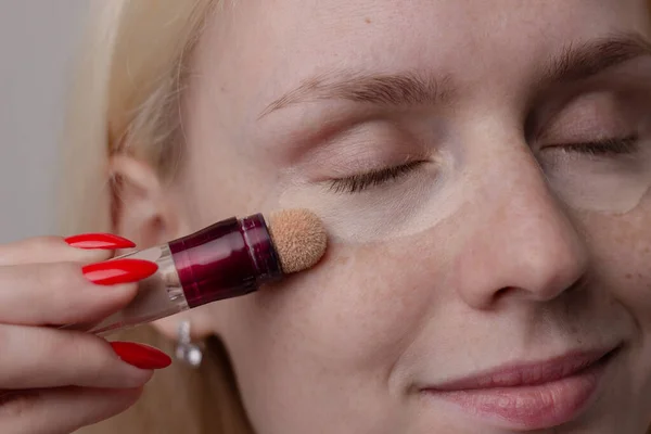  A young blonde woman with freckles applies concealer on her face under her eyes and masks circles under her eyes. Natural makeup. Text