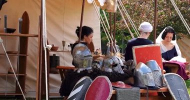 Kızlar yeni ortaçağ kıyafetlerini askeri çadırın ve ortaçağ savaş malzemelerinin yanına dikiyorlar.