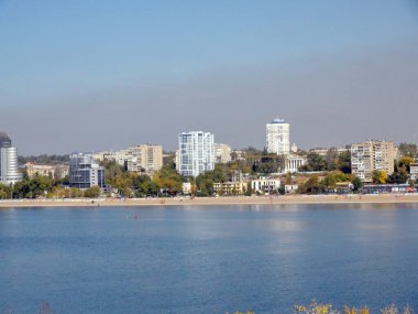 Landscape of the city beach near the blue mirror surface of the Dnieper against the background of skyscrapers shrouded in industrial emissions.