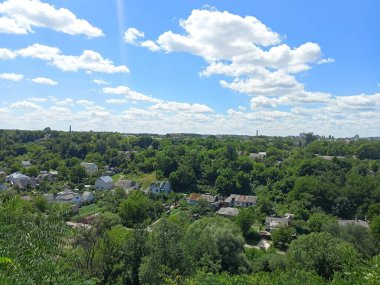 A bird's-eye view of the outskirts of the city drowning in the green of summer nature against the background of a sunny blue semi-cloudy sky.
