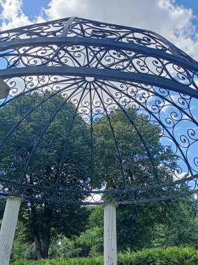 Bottom view while relaxing on a park decorative gazebo surrounded by tall green trees under a cloudy blue sky.