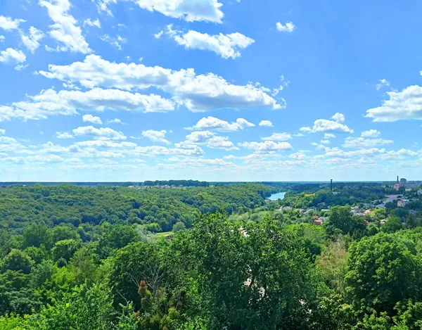 A panorama from the observation wheel of Ukrainian forests cut by a silver thread of a small river against a background of sunny blue covered with snow-white clouds.