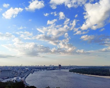A fantastic panorama of the cloudy sky at sunset that illuminates snow-white clouds with iridescent colors over the mighty Dnieper river bound by Kyiv bridges.