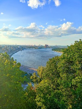 An unparalleled bird's-eye view of the rapid current of the Dnieper surrounded by the summer greenery of Kyiv streets against the background of the blue evening sky on the horizon.
