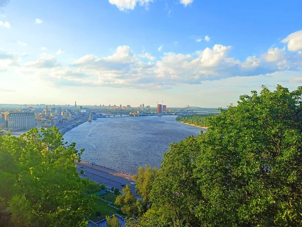 An unparalleled bird\'s-eye view of the rapid current of the Dnieper surrounded by the summer greenery of Kyiv streets against the background of the blue evening sky on the horizon.