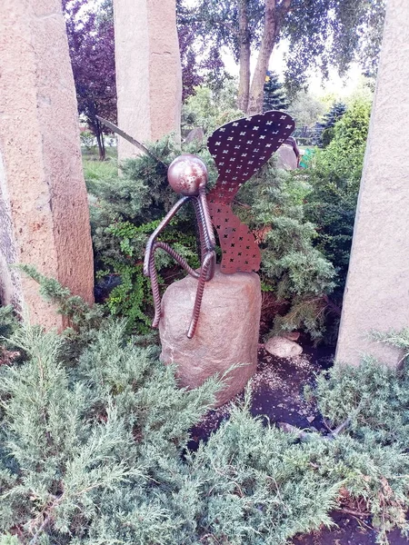 A metal sculpture of a fairy-tale butterfly that tiredly sat down on a stone with its legs hanging down after an exhausting long flight over park flowers.