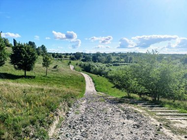 A wide dirt road in the middle of the steppe that bypasses a green hill and stretches to the cloudy sky on the horizon.