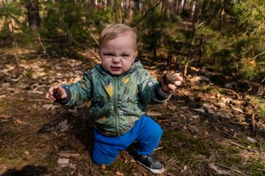 little boy with blond hair in the forest holding a pine cone