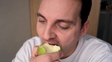 a man in a white sweater bites an apple looks pensively into the distance