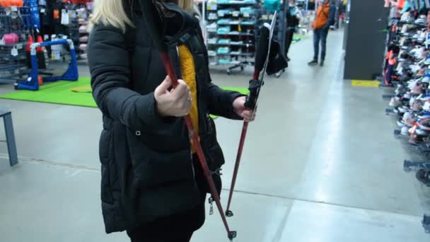 Μια νεαρή γυναίκα επιλέγει κοντάρια για σκανδιναβικό περπάτημα σε ένα κατάστημα αθλητικών ειδών — Αρχείο Βίντεο