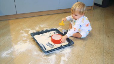 Küçük bir çocuk mutfakta unla oynuyor. Un ve küreği kum havuzunda gibi alıyor.