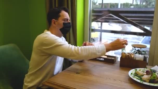 Egy férfi leül egy asztalhoz egy étteremben, maszkot visel, vesz egy fertőtlenítőt és ráfröcsköli a kezére. — Stock videók