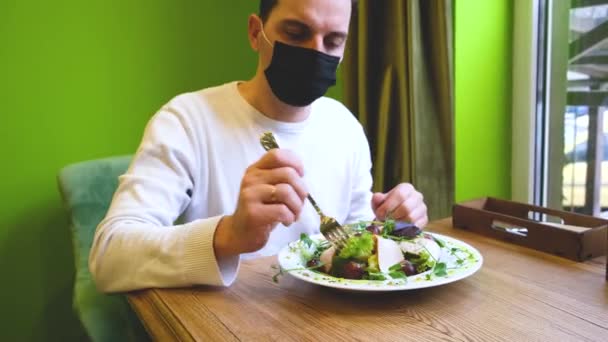 Un hombre se sienta en una mesa en un restaurante con una máscara, baja la máscara pone comida en su boca y pone la máscara de nuevo — Vídeo de stock